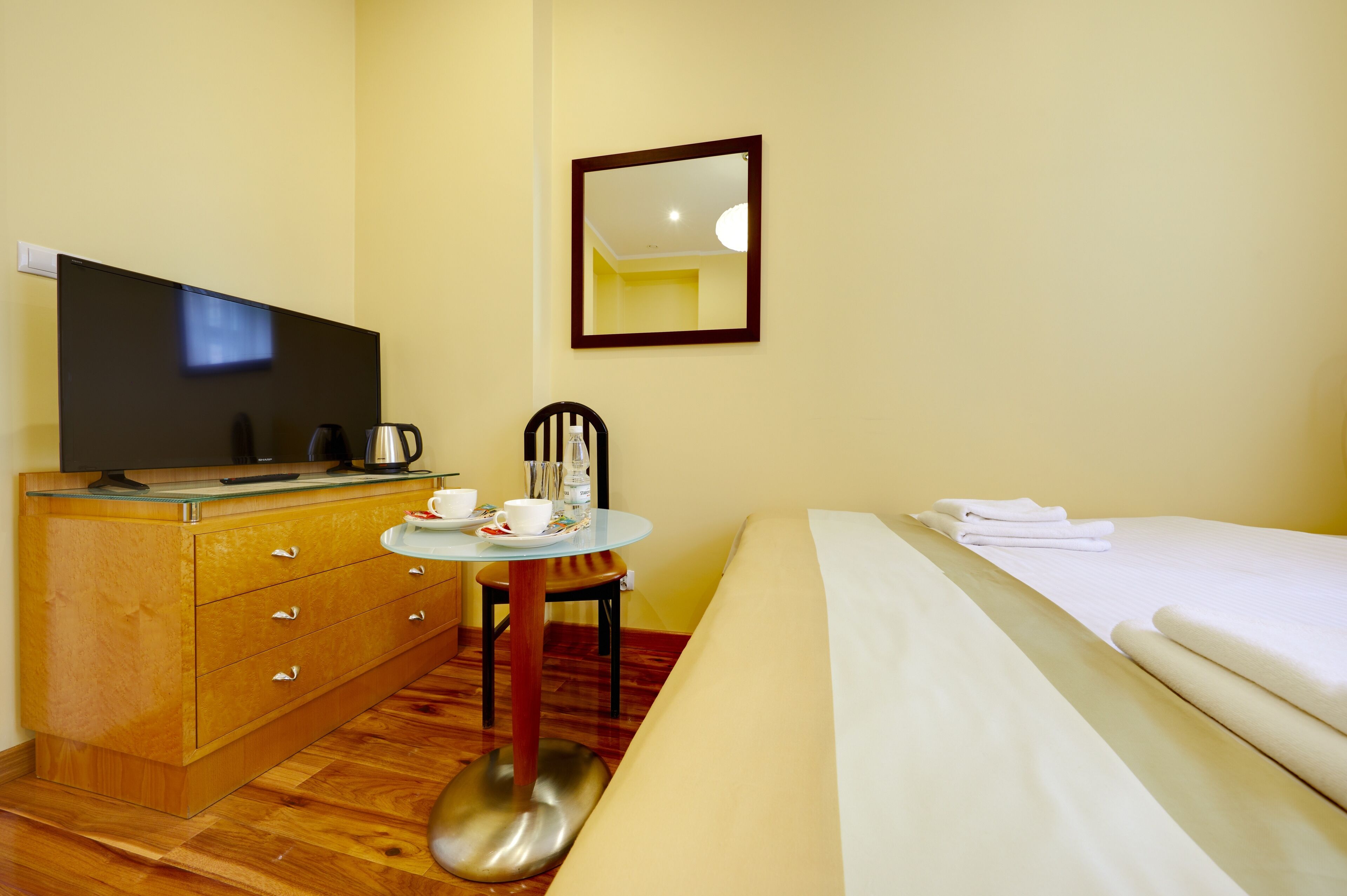 A hotel room with a wooden dresser, a television, a small round table with a chair, a bed with white linens, and a mirror on the wall.