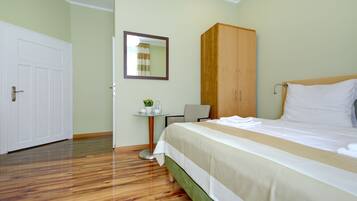 Habitación de hotel con piso de madera, cama con ropa blanca, armario de madera, una mesita con una planta y un espejo en la pared.