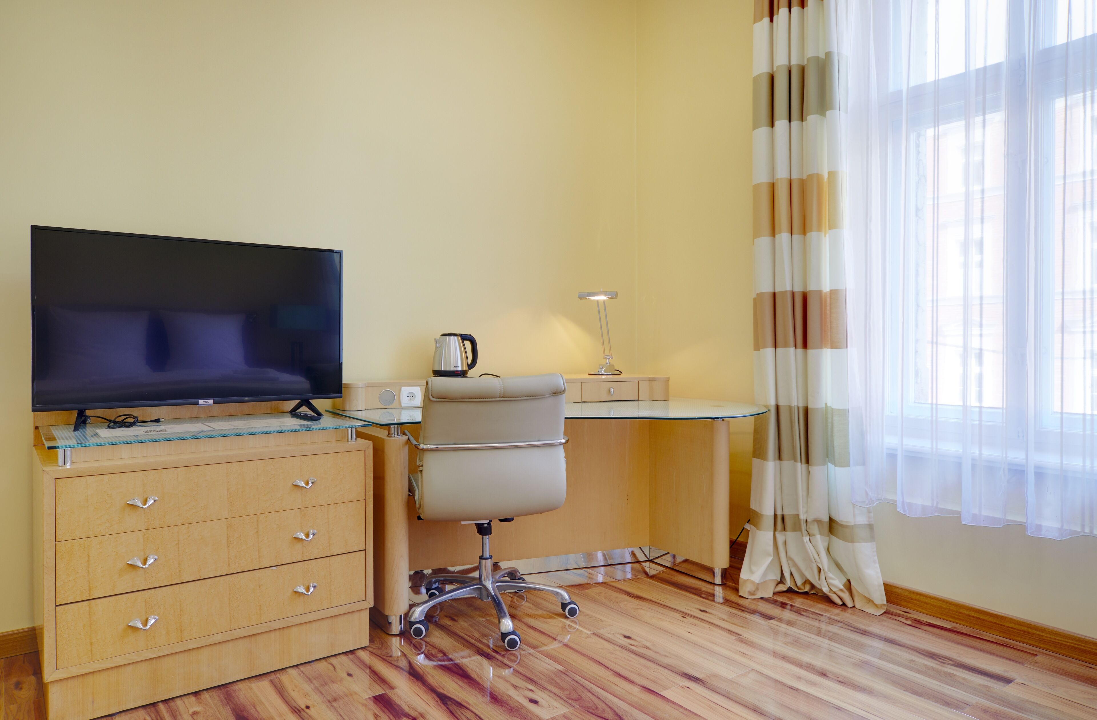 A hotel room with a wooden desk, a television, a chair, a lamp, and a window with curtains.