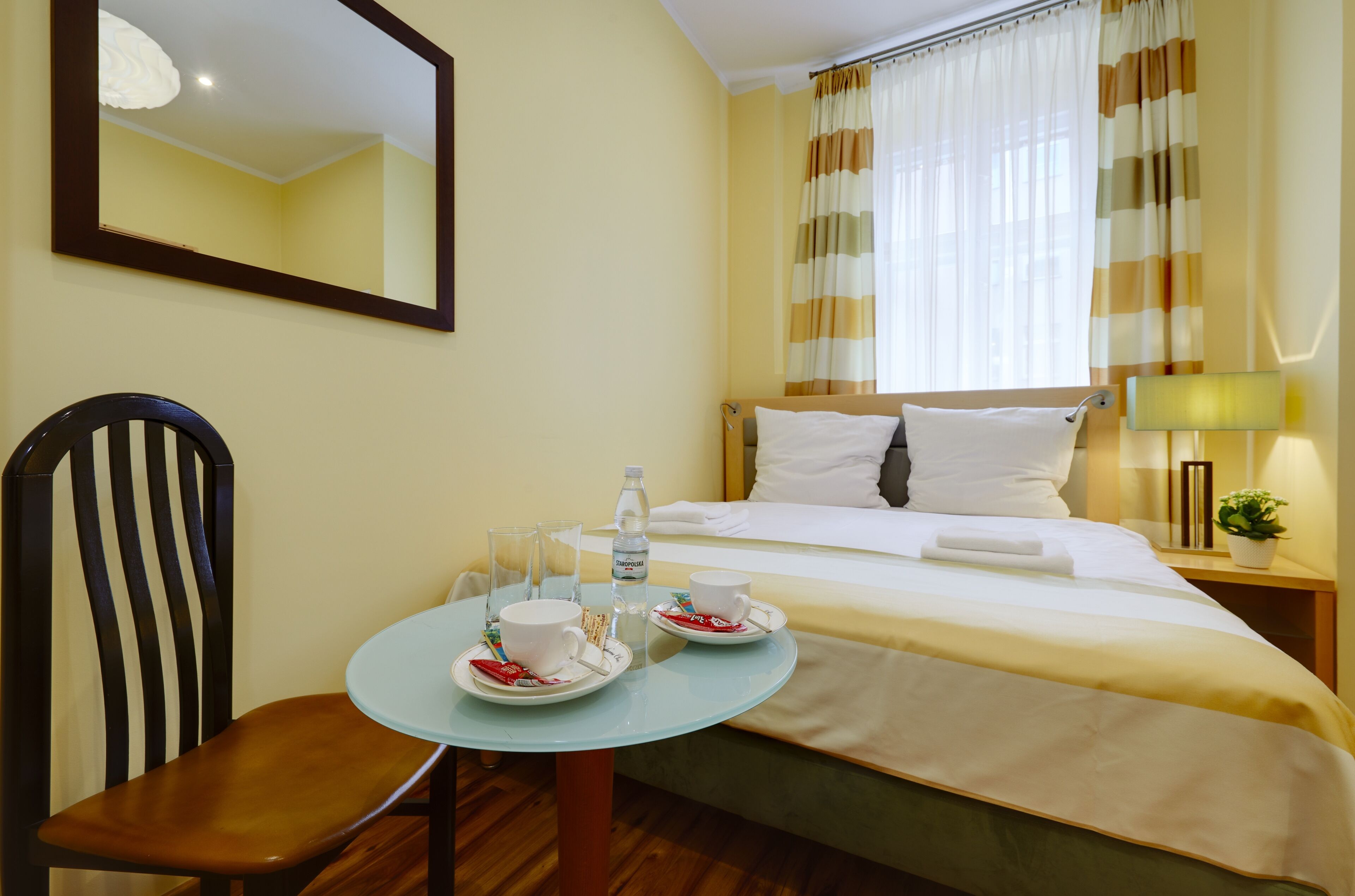 A hotel room with a bed, a bedside table with a lamp, a chair, and a small table with two cups and a bottle.