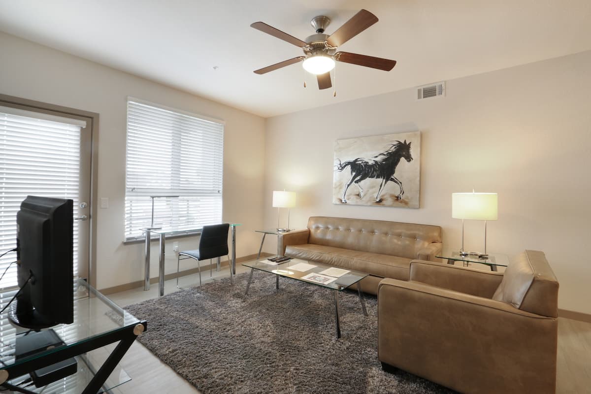 A living room with a brown leather sofa, a glass coffee table, a television on a glass stand, two lamps, and a ceiling fan.