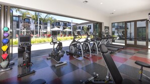 Un gimnasio moderno con diversos equipos de ejercicio, ventanas amplias y vistas a edificios y zonas verdes.