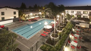Una piscina con sillones y mesas, rodeada de edificios y follaje.