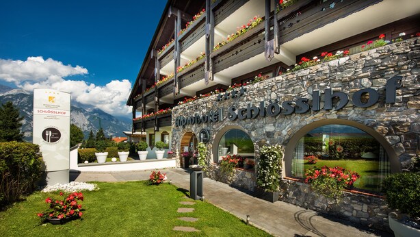 Un hotel con fachada de piedra, balcones con jardineras florales y una vista despejada de montañas al fondo.