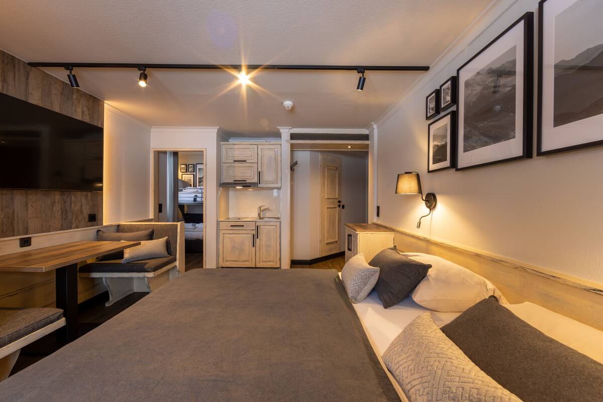 A modern bedroom with a large bed, bedside tables, a desk, and a kitchenette.