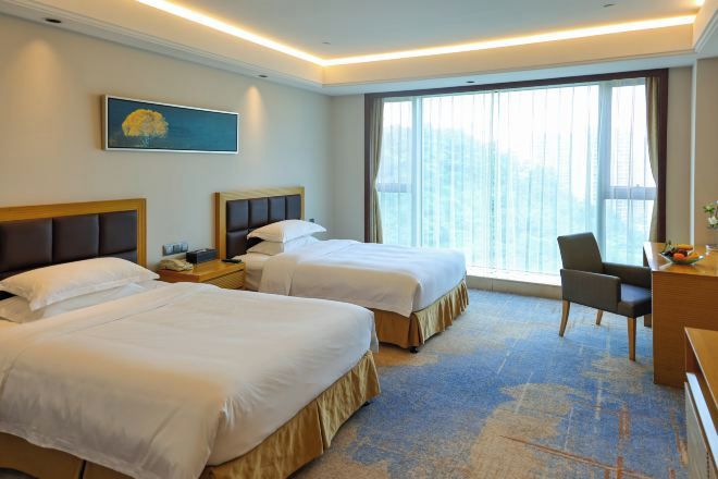 A hotel room with two beds, a desk, a chair, a large window with curtains, and a painting on the wall.