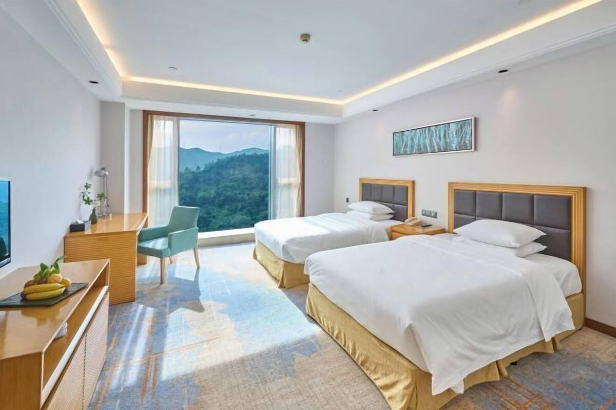 A hotel room with two beds, a desk, a chair, and a large window with a mountain view.