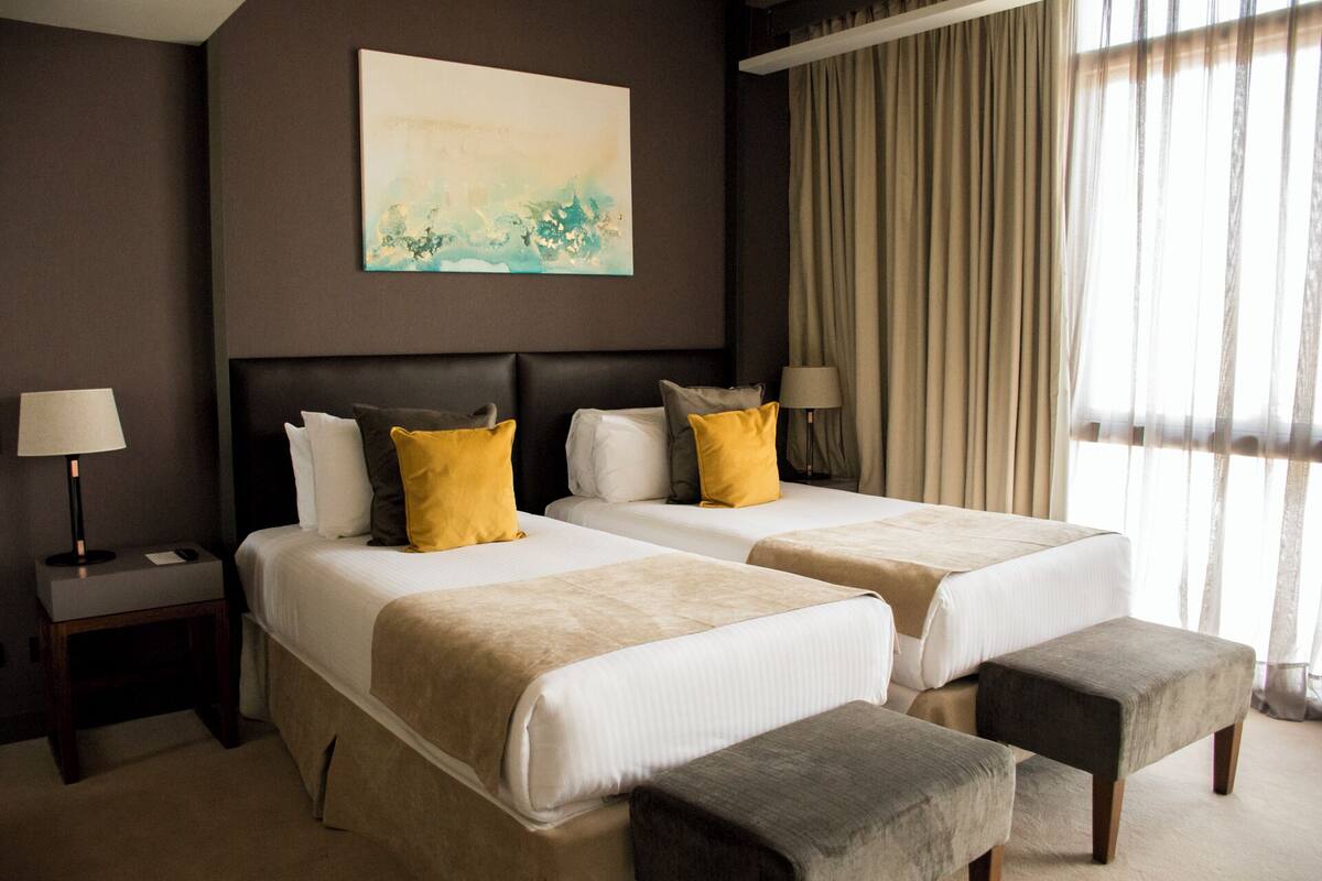 Une chambre d’hôtel avec deux lits, une table de chevet, une lampe, un tableau et une fenêtre avec des rideaux.