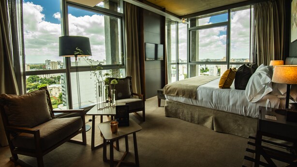 Een moderne hotelkamer met een groot bed, een bureau, een stoel en uitzicht op de stad door grote ramen.