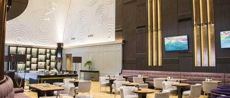 A spacious lobby with a modern design, featuring a reception area, seating arrangements, and a large screen displaying a sports event.