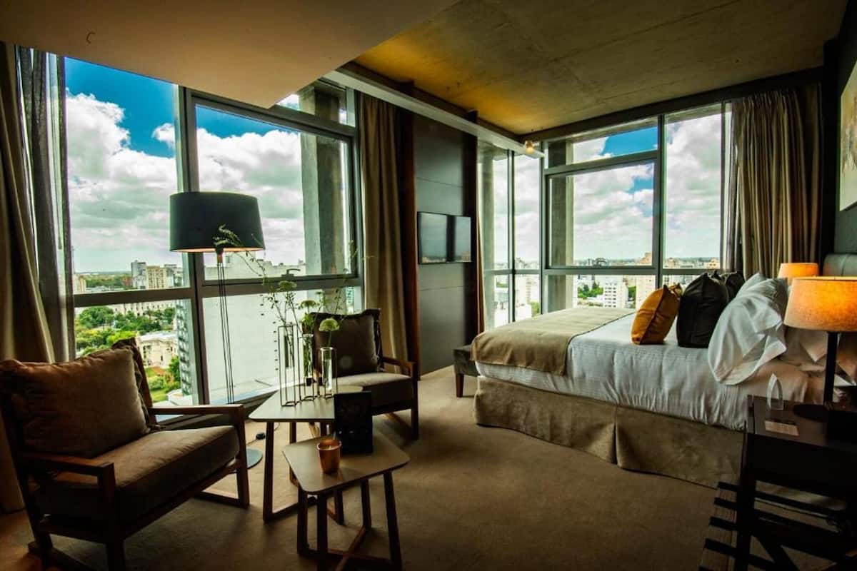 Une chambre d’hôtel avec un grand lit, un bureau, une chaise et une vue sur la ville grâce à de grandes fenêtres.