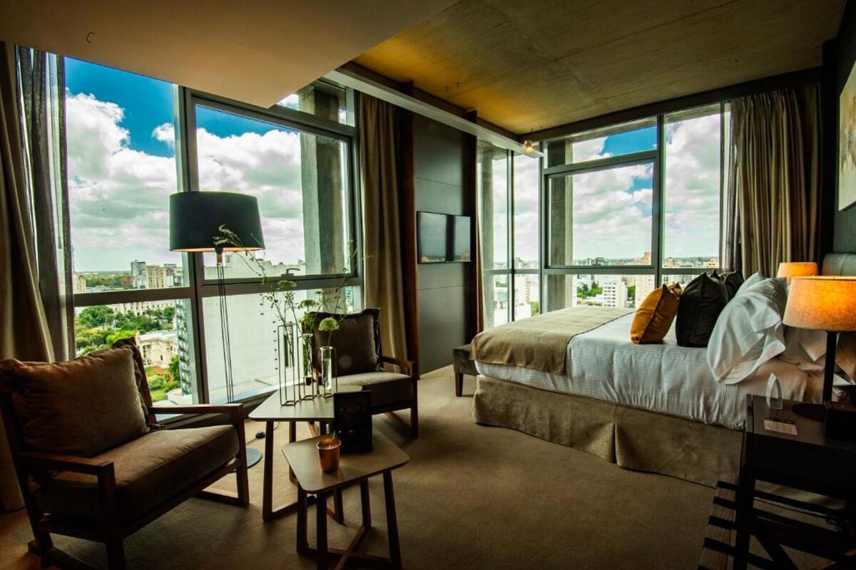 A hotel room with a large bed, a desk, a chair, and a view of the city through large windows.