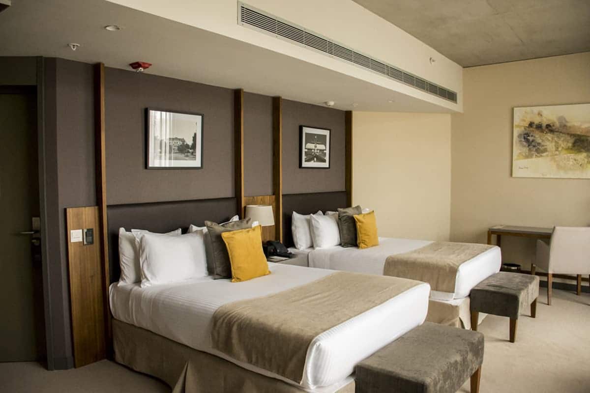 Une chambre d’hôtel avec deux lits, un bureau et une chaise. Les murs sont ornés de tableaux encadrés, et un tableau est accroché au-dessus du bureau.