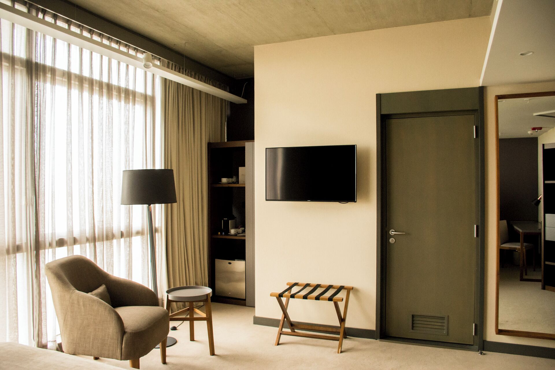 Habitación de hotel con sofá, una mesita redonda pequeña, una banca, televisor montado en la pared, una puerta y una ventana con cortinas.