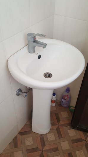 A bathroom with a white pedestal sink, a silver faucet, and a drain. There is a bottle of cleaning product and a green basket on the floor.