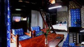 A room with a wooden table, blue patterned chairs, a bed with white bedding, and a stone wall. - Treetops Lodge (Port Vila)