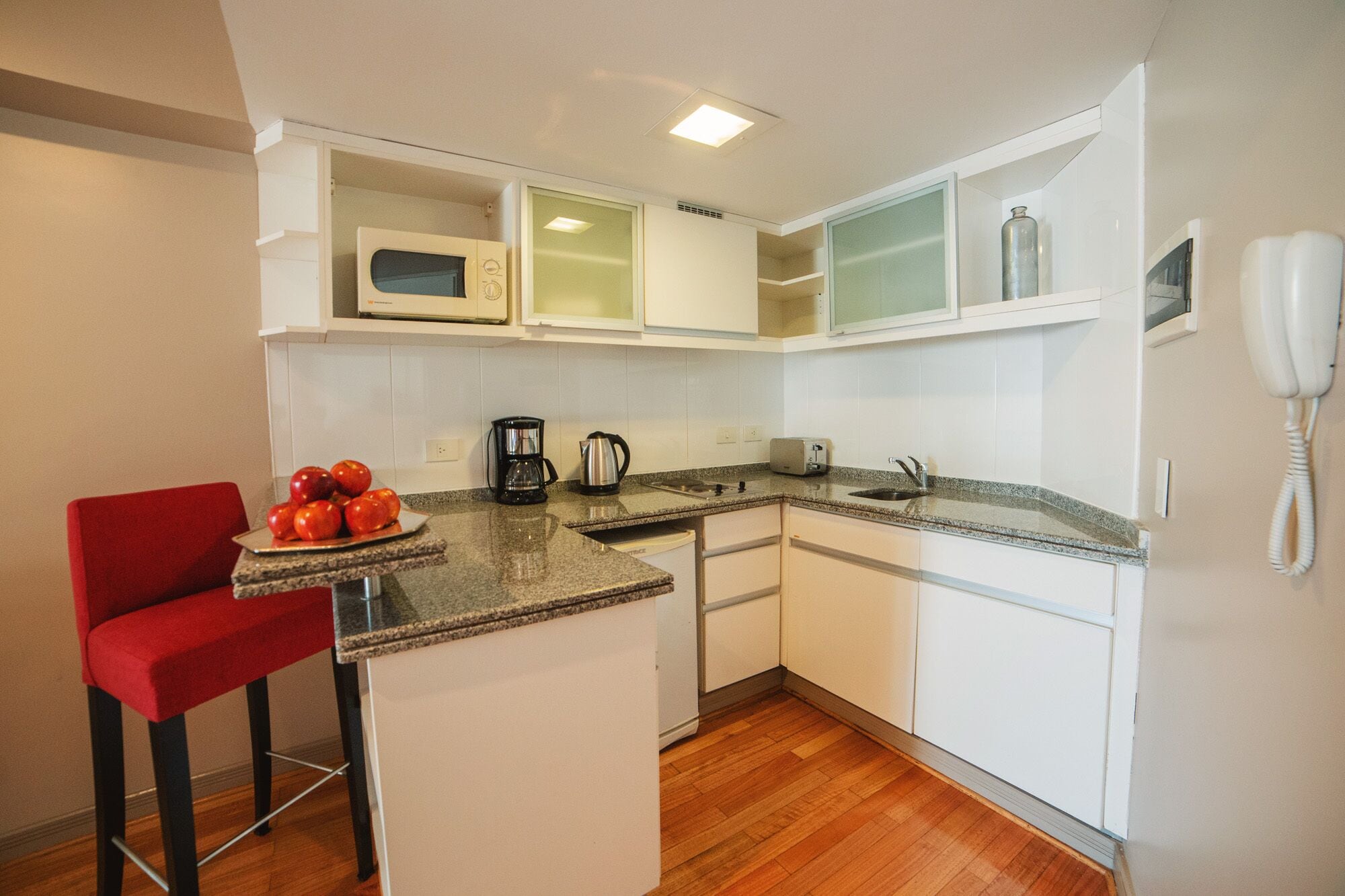 A compact kitchen with white cabinets, a microwave, a coffee maker, and a toaster. There is a red chair and a plate of apples on the counter.
