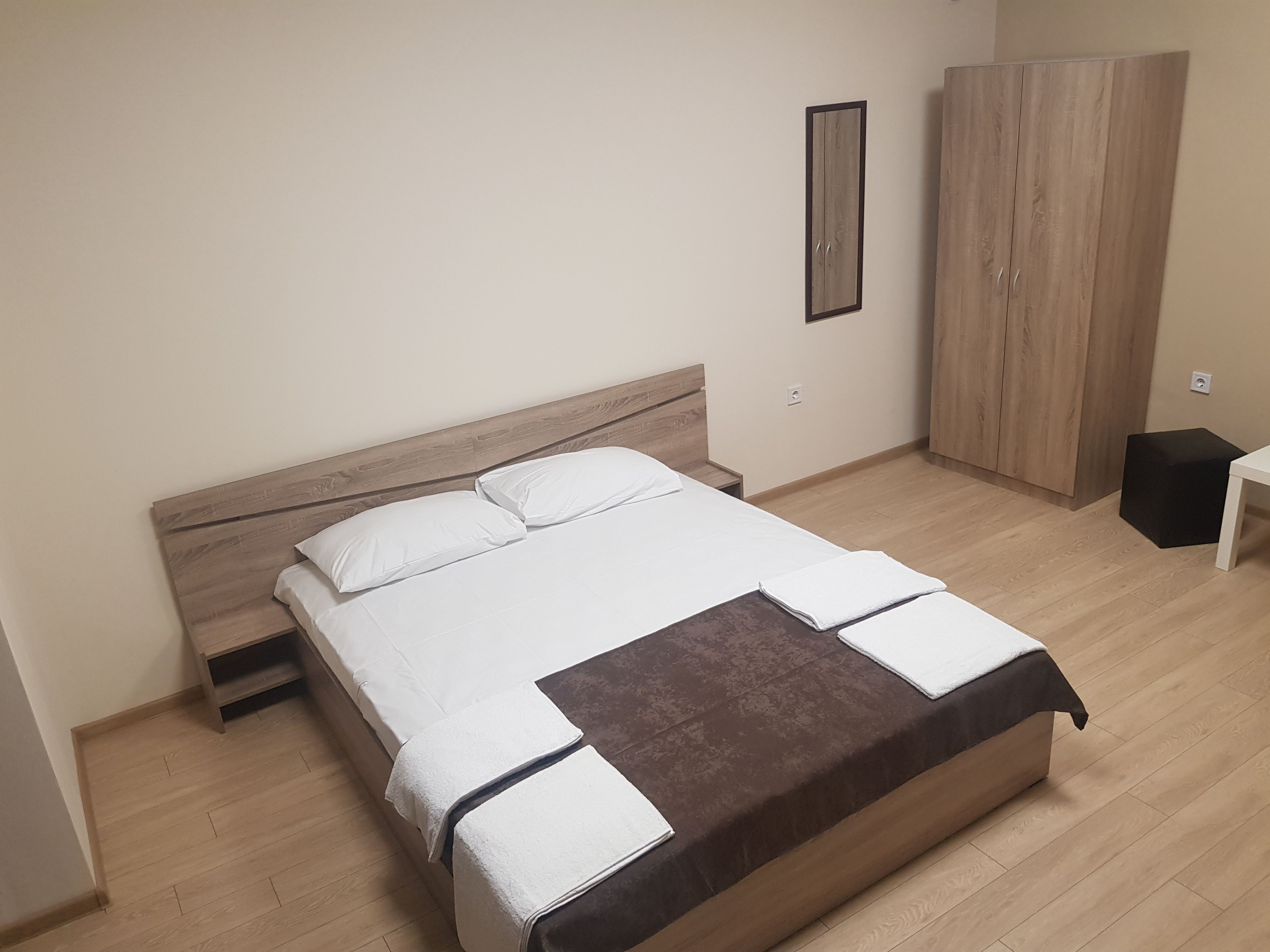 Una cama bien tendida con sábanas blancas y una manta marrón, un armario de madera y una mesita de noche con una lámpara.