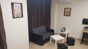 Ein modernes Wohnzimmer mit einem grauen Sofa, einem weißen Couchtisch, einem schwarzen Hocker, einem kleinen weißen Beistelltisch mit Wasserkocher und einem Fernseher auf einem Holzgestell.
