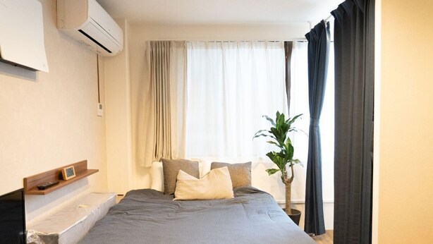 A hotel room with a bed, a bedside table, a plant, and a window with curtains. - East Shinagawa Apartment (Tokyo)