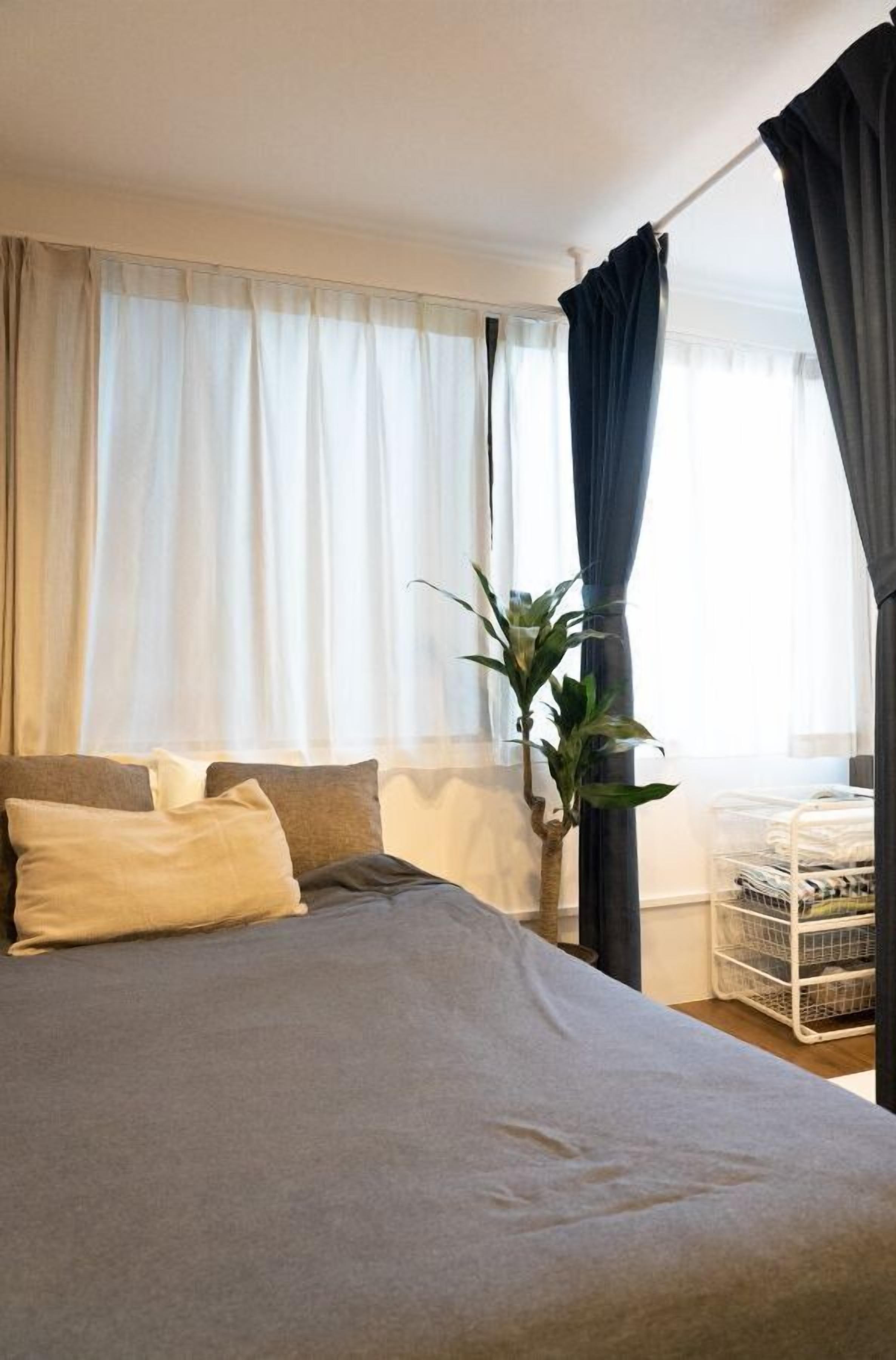 A neatly made bed with a grey bedspread and two pillows, a potted plant, and a white storage unit against a window with sheer curtains.