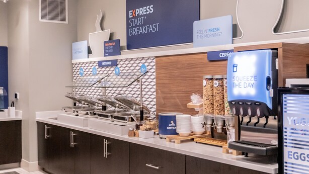 A breakfast buffet area with labeled food stations, a coffee machine, and a sign indicating 'Express Start Breakfast'.