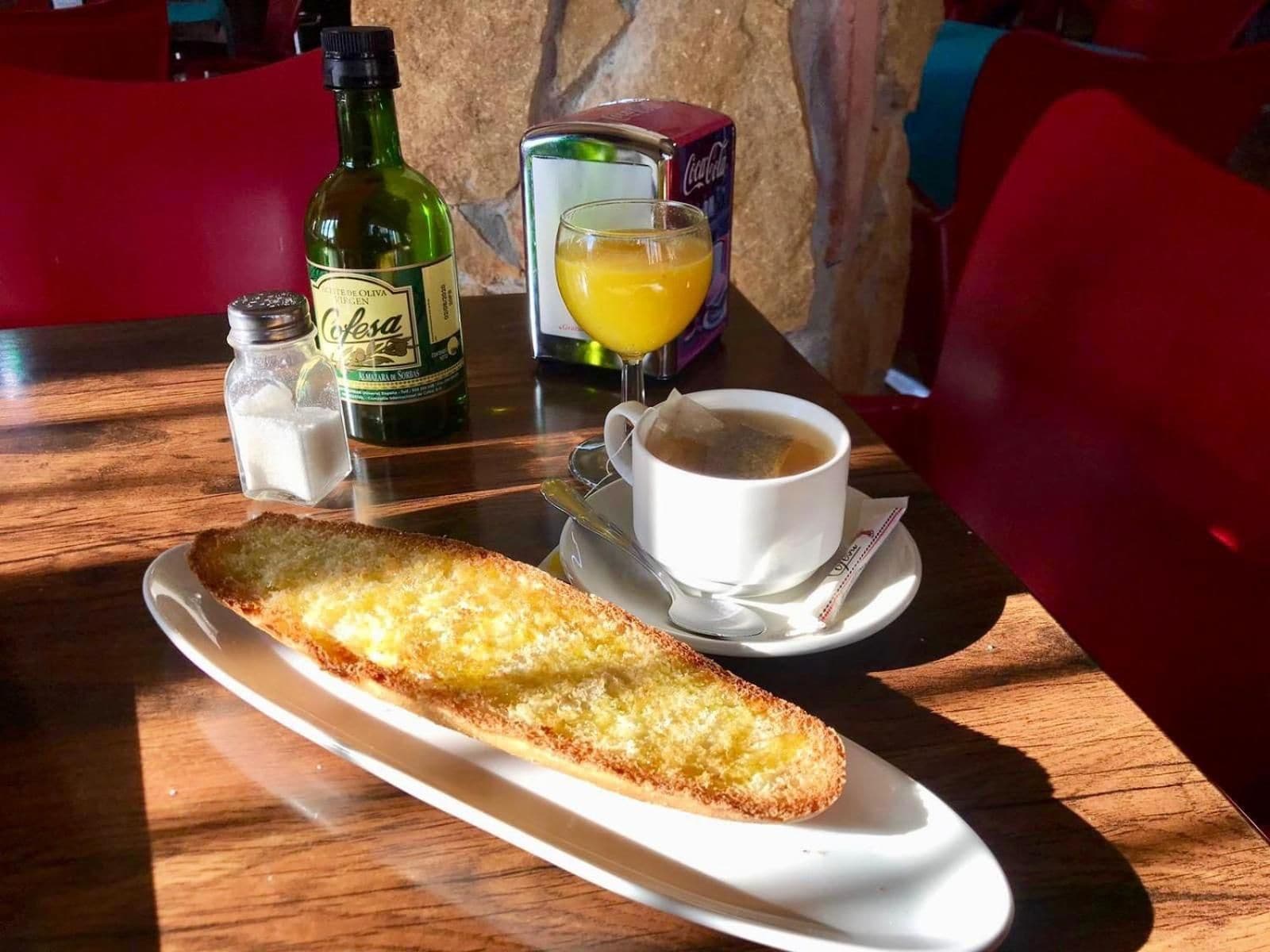 A table with a plate of bread, a cup of coffee, a glass of orange juice, a bottle of olive oil, and a salt shaker.