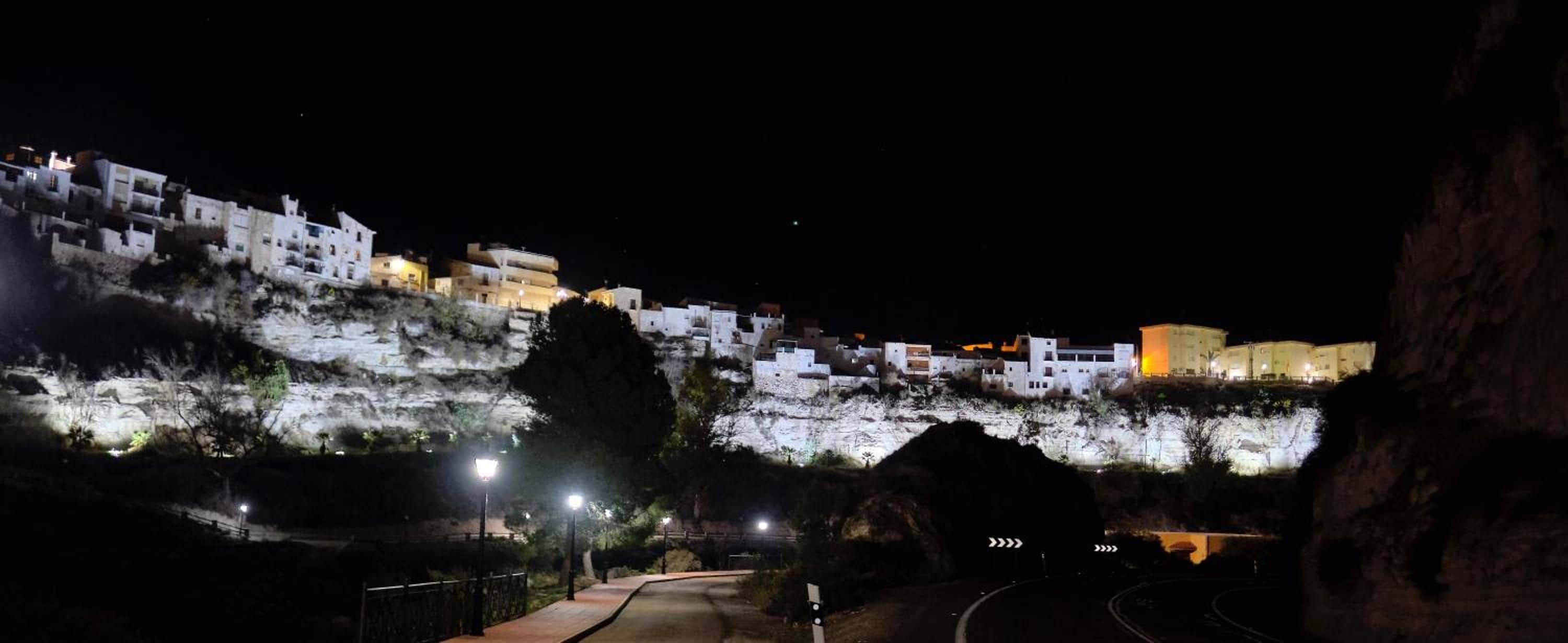 A nighttime view of a hillside with illuminated buildings, a paved pathway, and a guardrail.