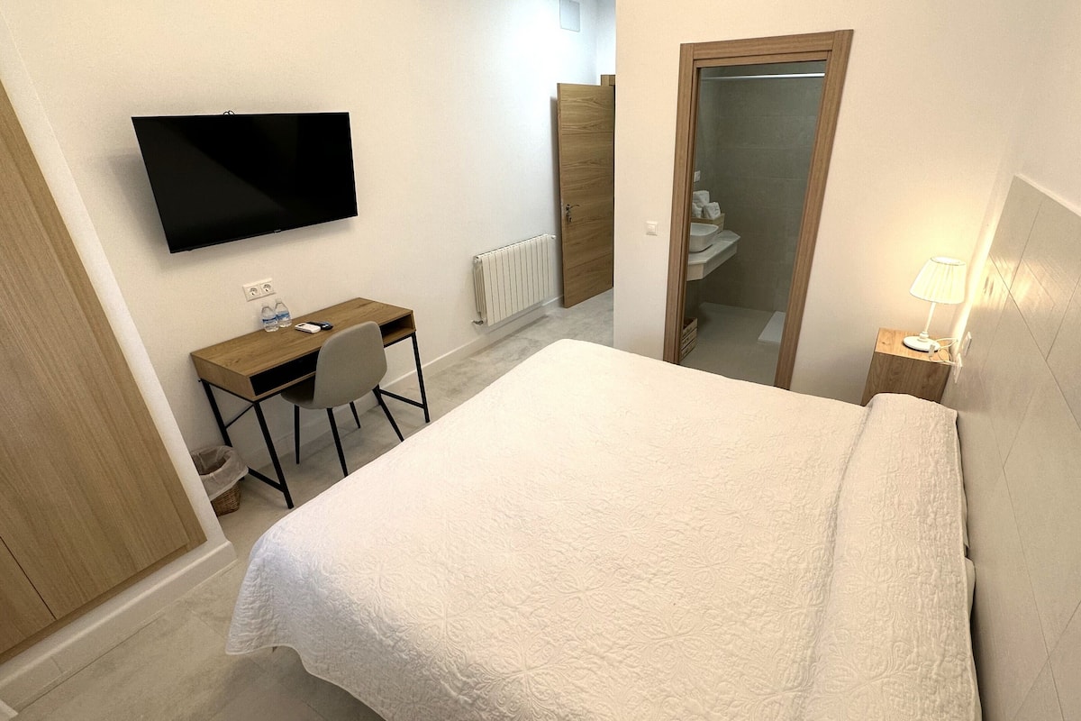 Una habitación de hotel con una cama, un escritorio con una silla, un televisor, un espejo y una mesita de noche con una lámpara.