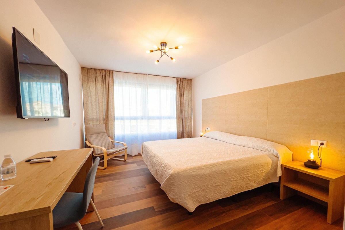 Habitación de hotel con cama, escritorio, silla, televisor y ventana con cortinas.