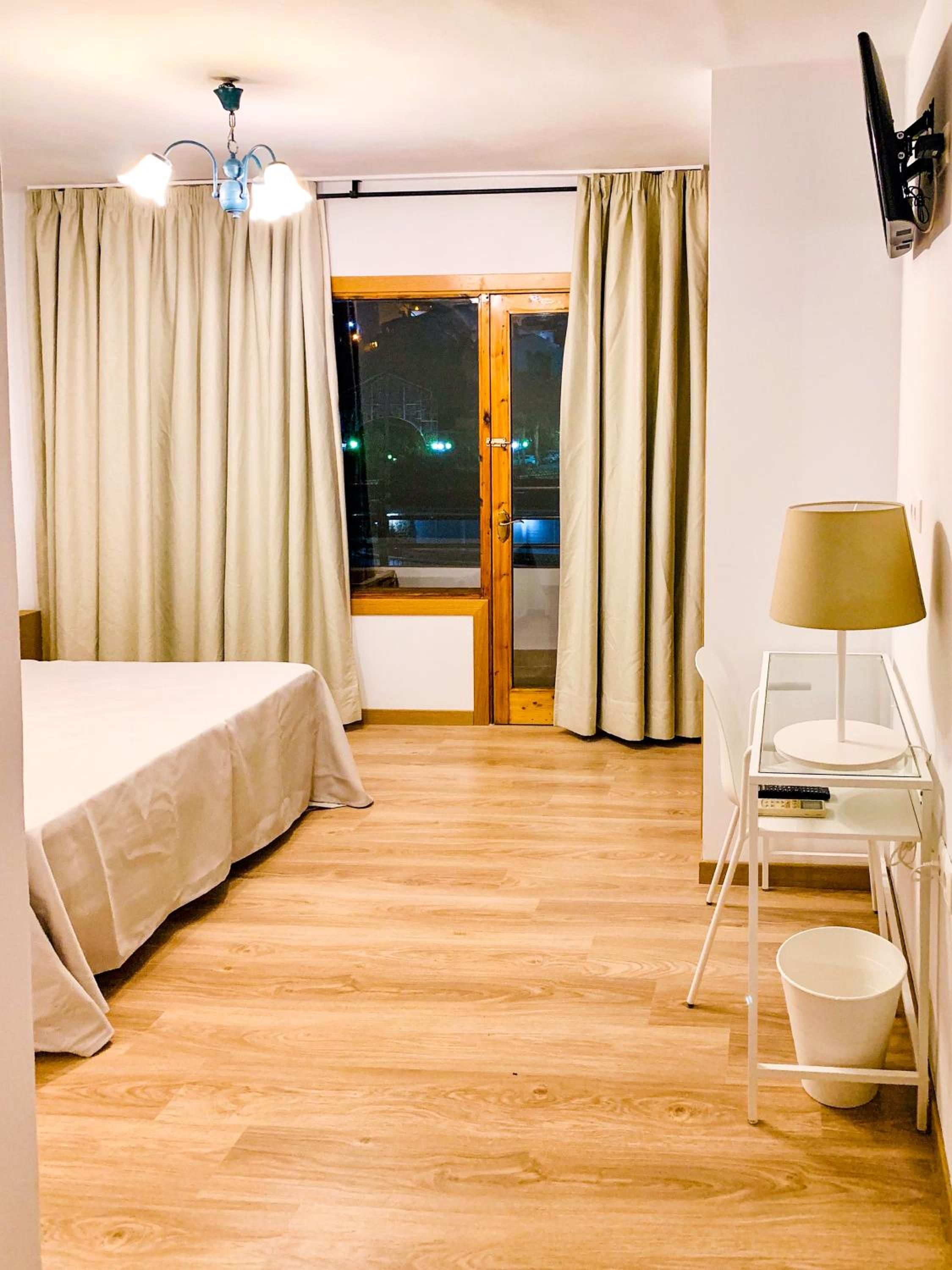 A hotel room with a bed, a desk with a lamp, a chair, a small table, and a view of the outside at night.