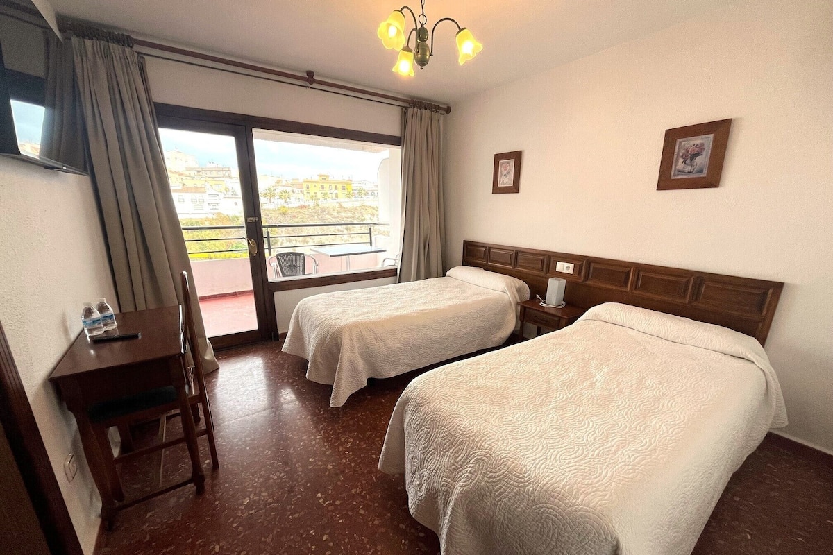 Habitación de hotel con dos camas, un escritorio, una silla, una mesita y un balcón con vistas.