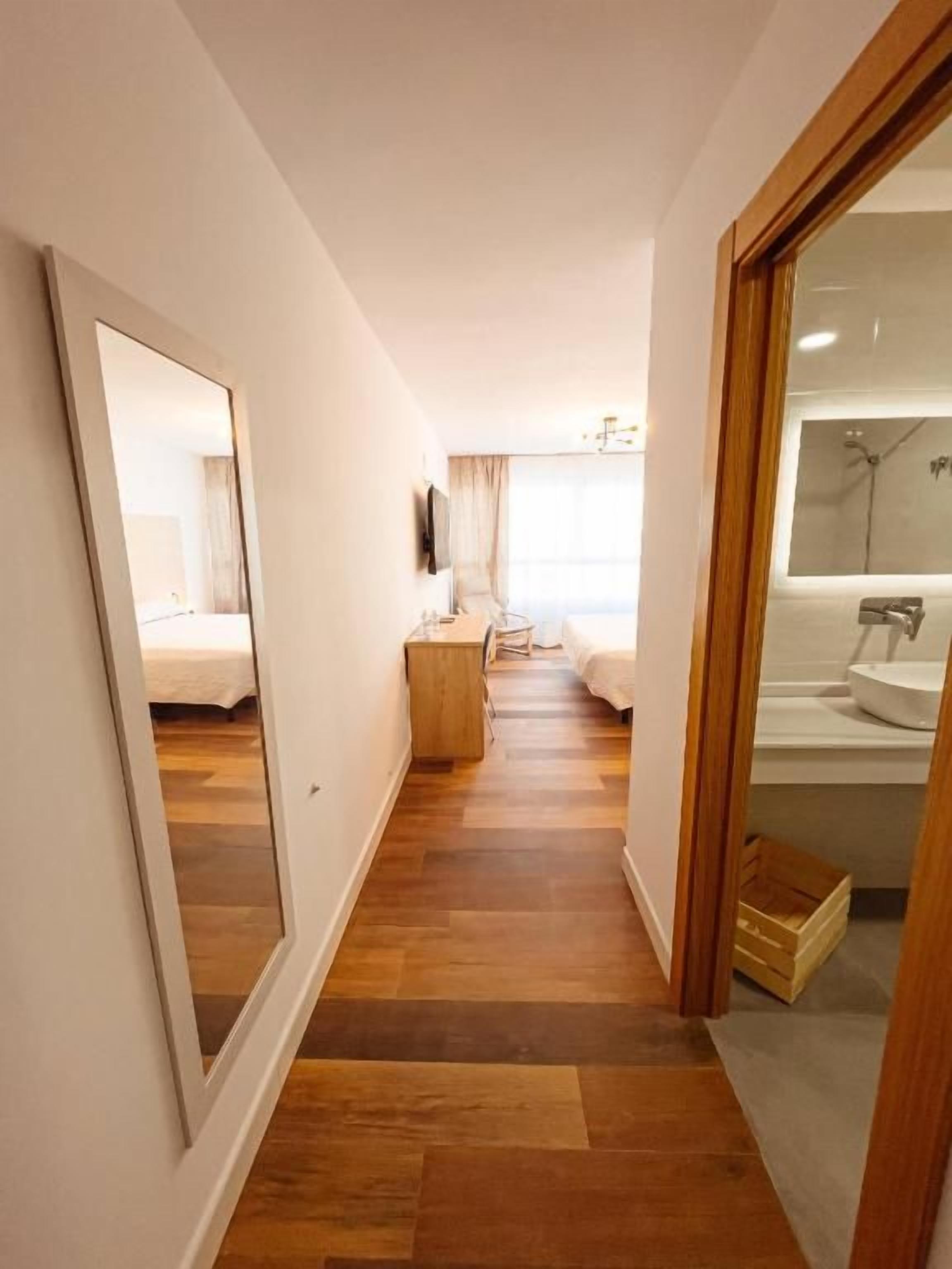 A hotel corridor with a wooden floor, a bed, a mirror, a television, and a bathroom with a sink and mirror.