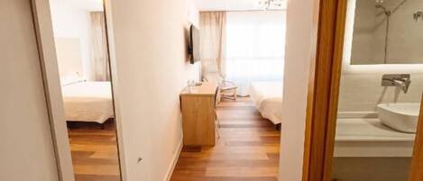 A hotel corridor with a wooden floor, a bed, a mirror, a television, and a bathroom with a sink and mirror.