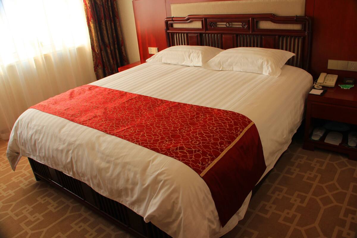Une chambre d’hôtel avec un grand lit, une tête de lit en bois, une table de chevet avec un téléphone et un sol recouvert de moquette.