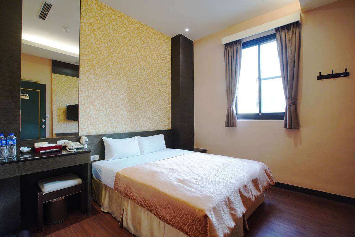 Habitación de hotel con cama, escritorio, teléfono, cesto de basura y ventana con cortinas.