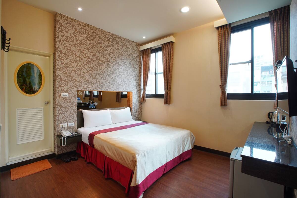 Habitación de hotel con cama, escritorio, ventana con cortinas, puerta con espejo y pared con estampado floral.