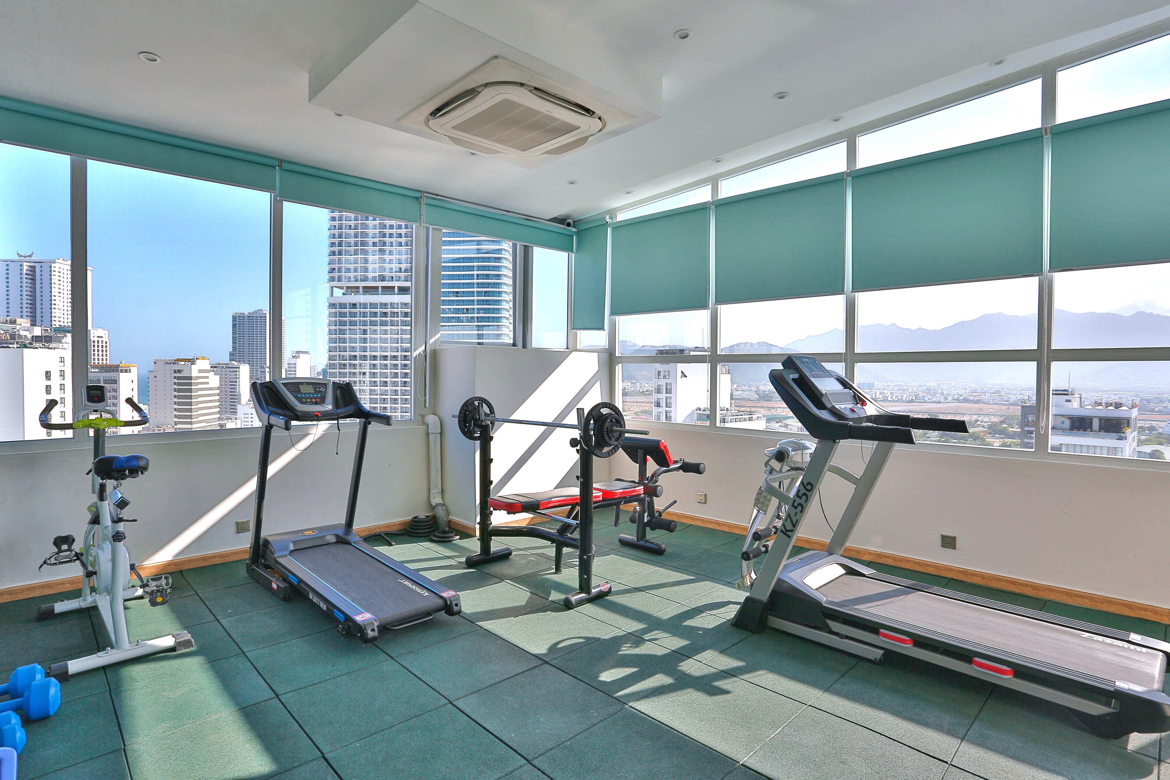 A gym with treadmills, a weight bench, and dumbbells, with a city view through large windows.