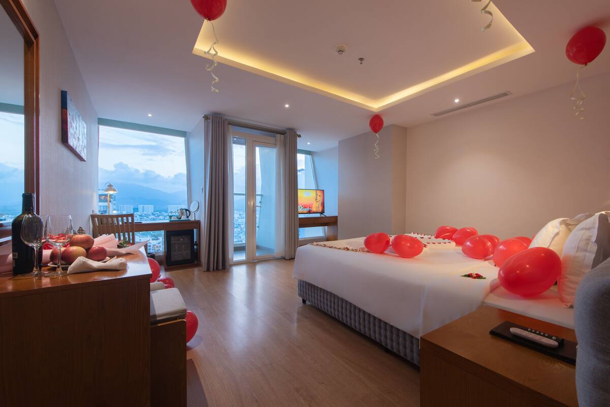 A hotel room with a bed, a desk with a laptop, a view of the city, and a balcony.