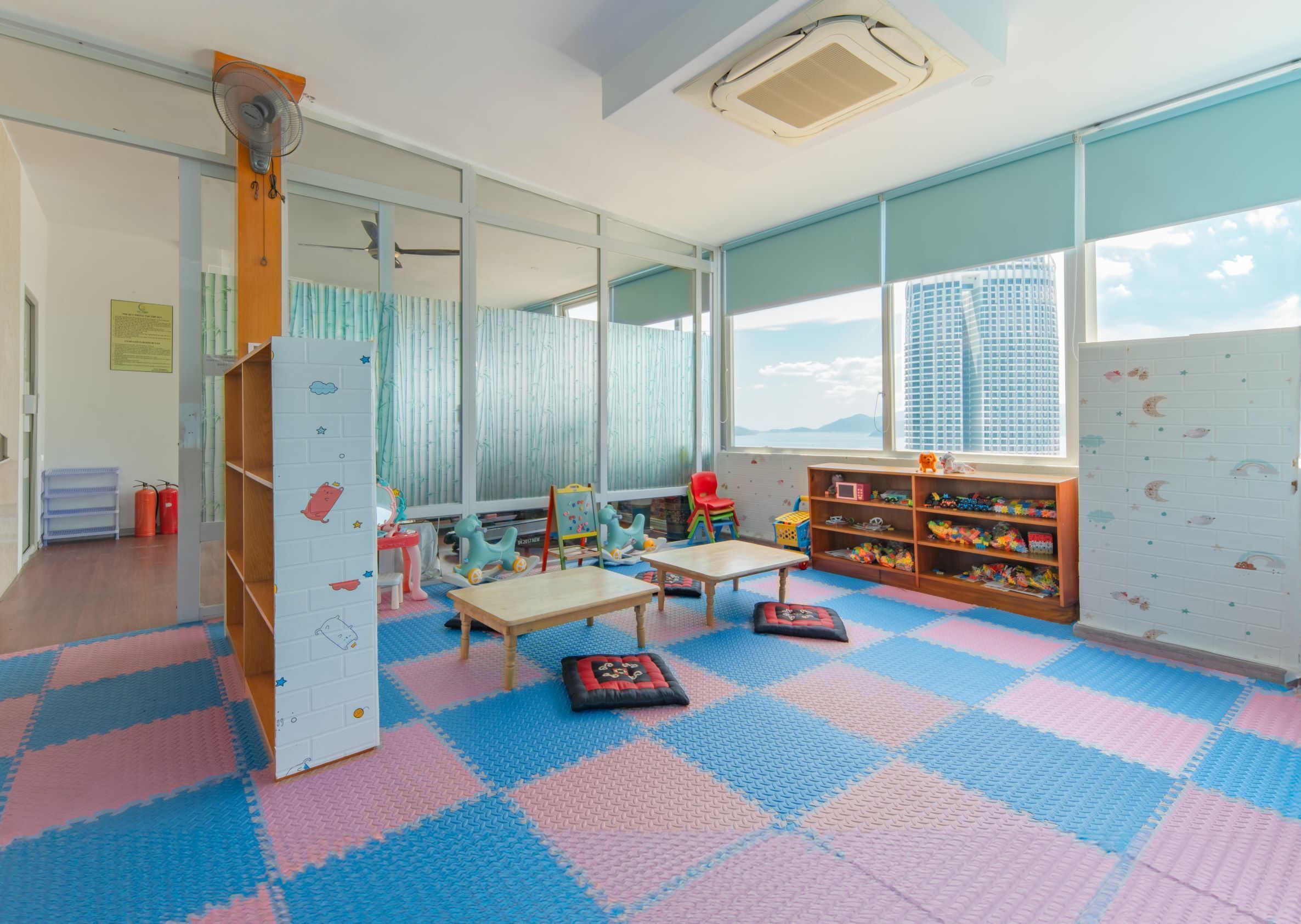 A children's playroom with a large window, a wooden play table, colorful toys, and a blue and pink checkered floor.