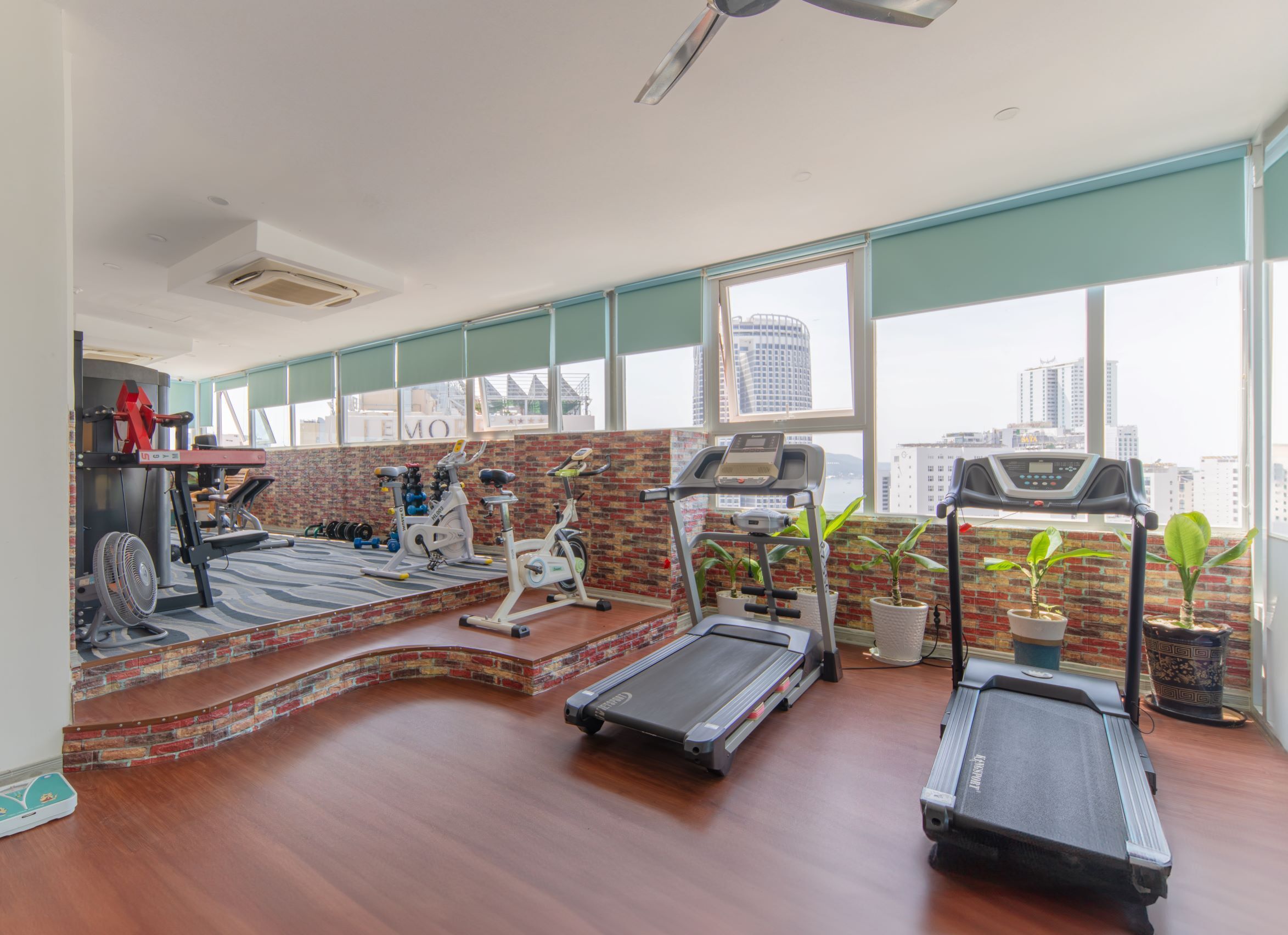 Ein Fitnessraum mit Laufbändern, einem Crosstrainer und einem Ventilator. Es gibt große Fenster und eine Ziegelwand.