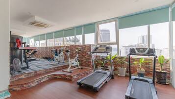 Ein Fitnessraum mit Laufbändern, einem Crosstrainer und einem Ventilator. Es gibt große Fenster und eine Ziegelwand.