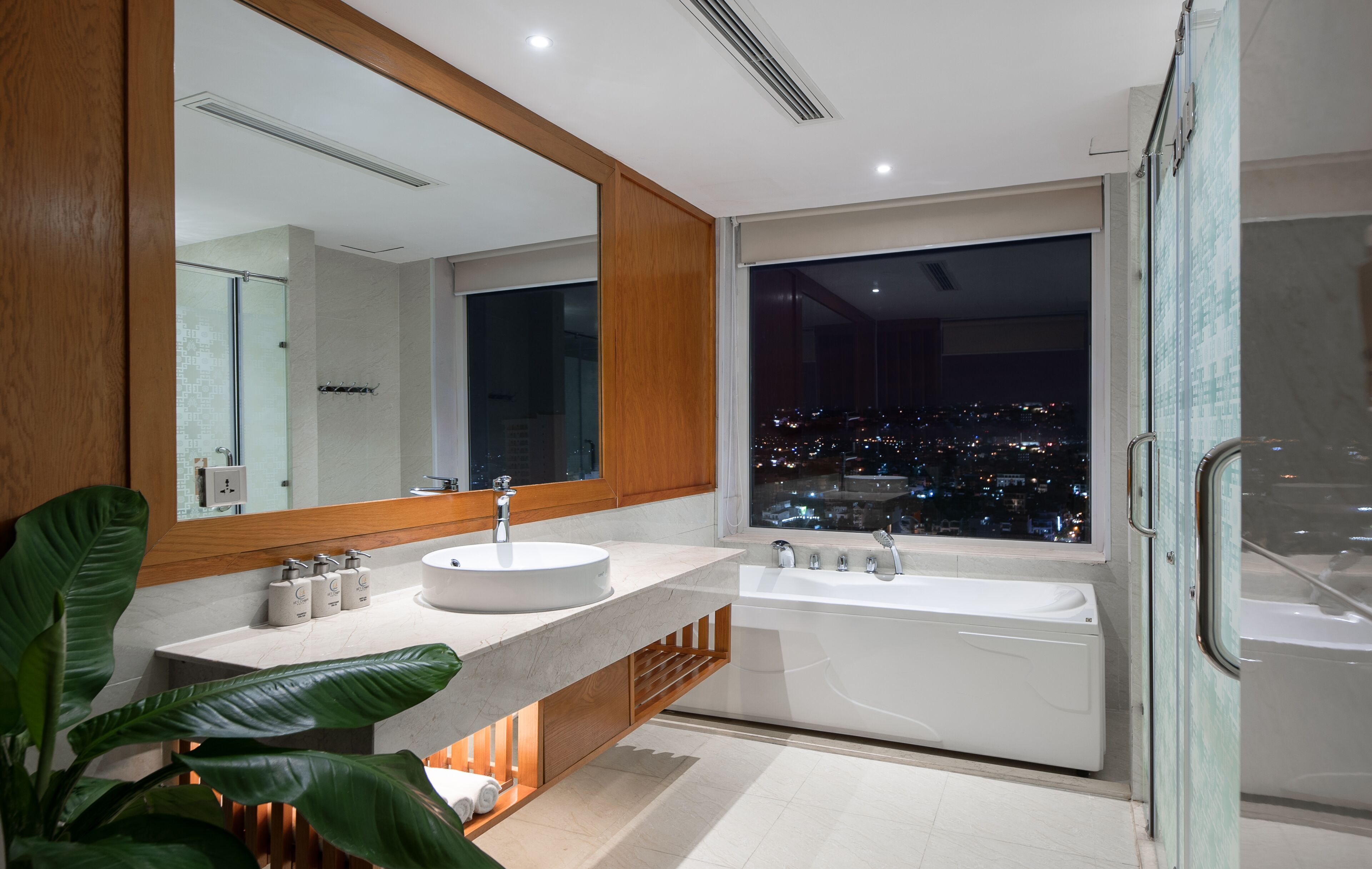 A modern bathroom with a large mirror, two sinks, a bathtub, and a city view.