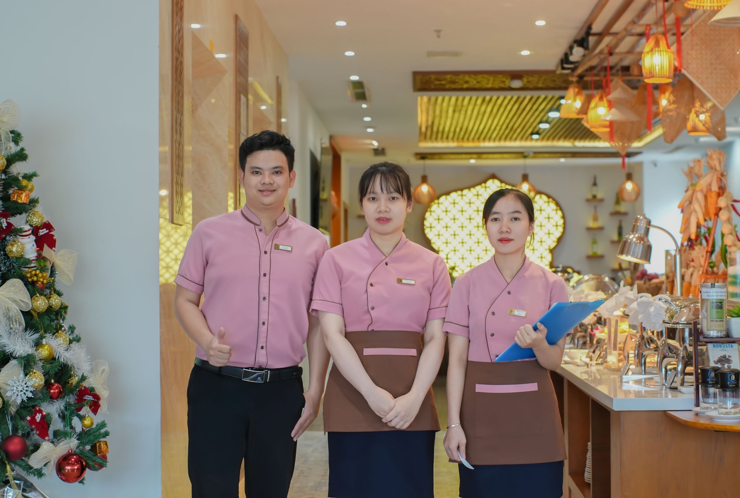 Three staff members in a hotel lobby with a Christmas tree and a reception desk.
