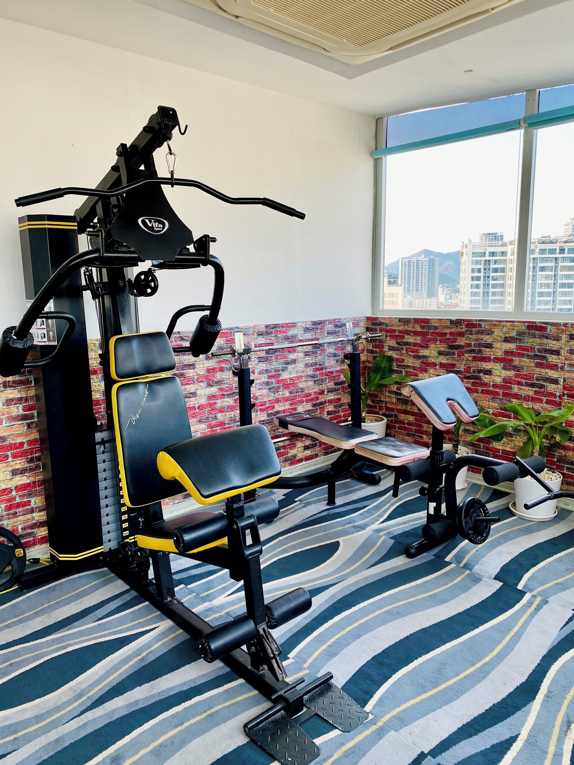 A gym with exercise equipment, a brick-patterned wall, and a view of buildings through large windows.