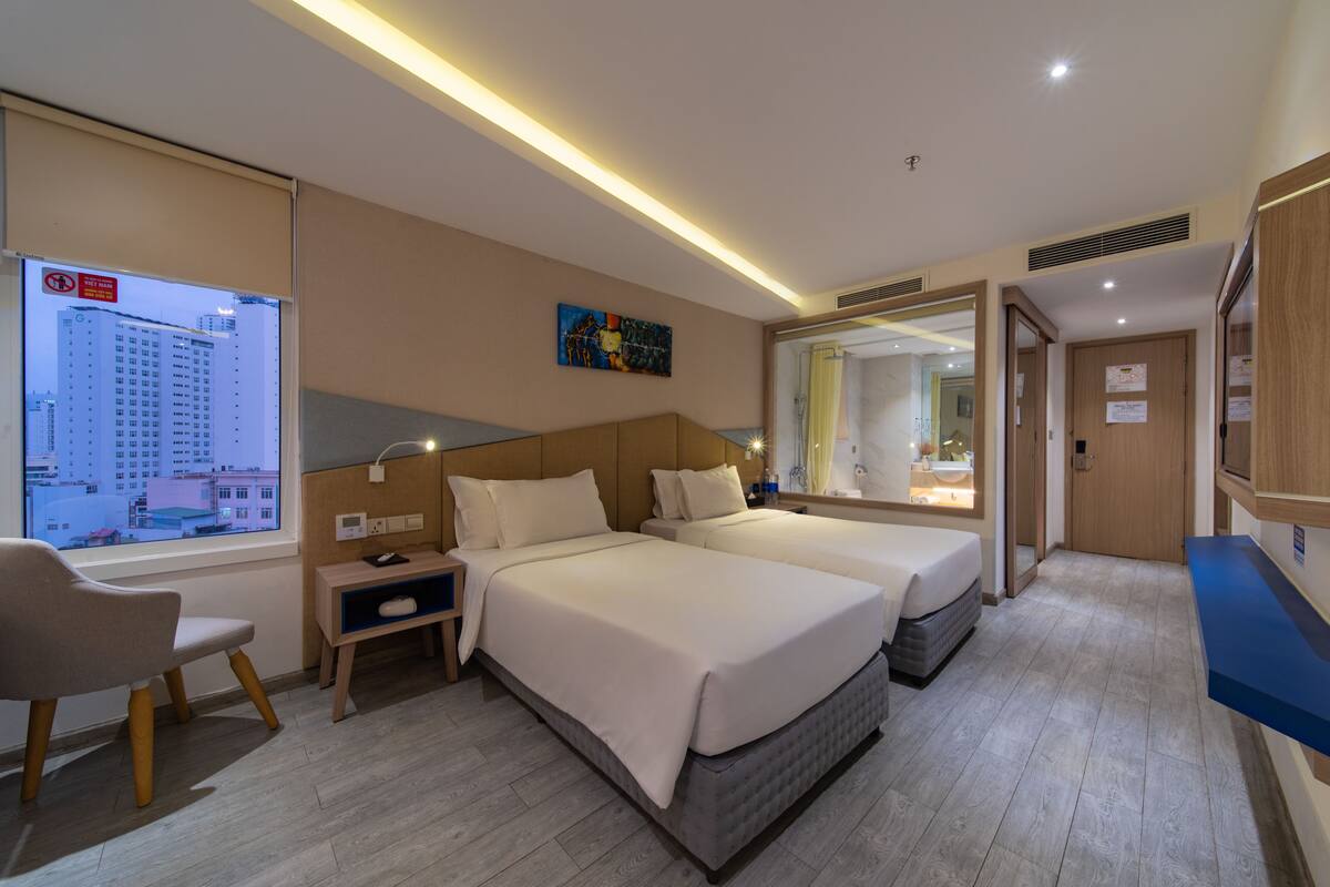 A hotel room with two beds, a desk, a chair, a large window, and a mirror.