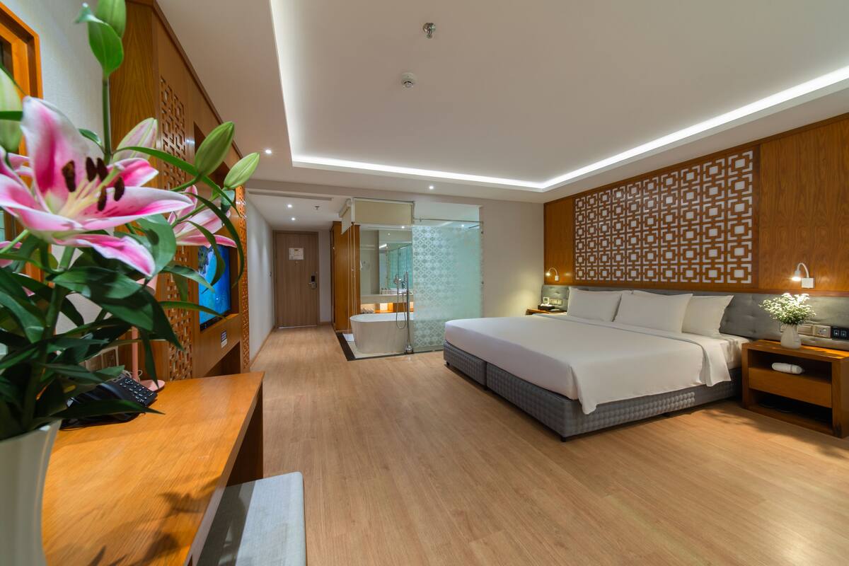 A modern hotel room with a large bed, wooden flooring, a desk with a computer, a bathroom with a glass shower, and a decorative wall panel.