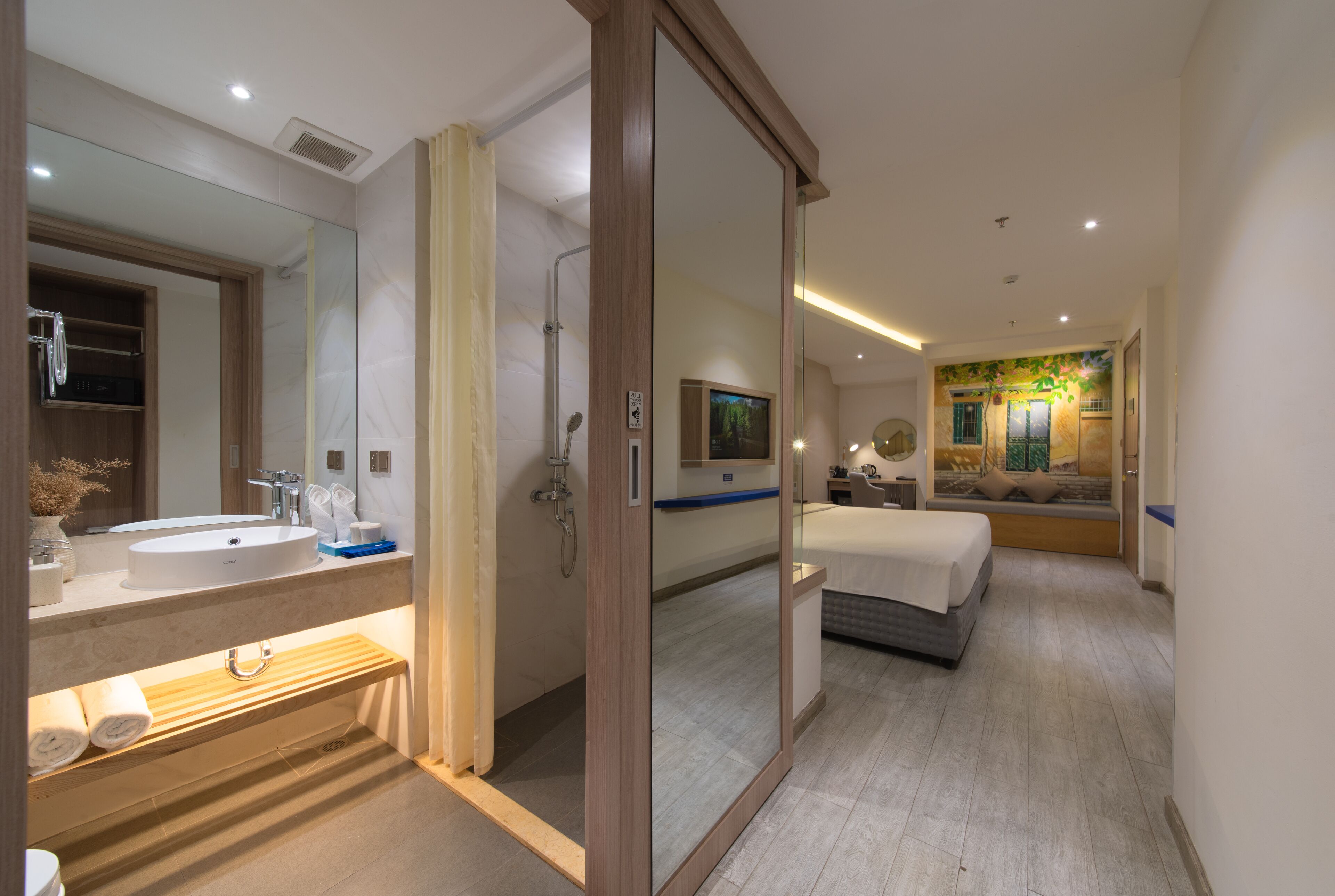 Un baño de hotel moderno con un espejo grande, lavamanos dobles, una ducha y una mesita de noche con lámpara.