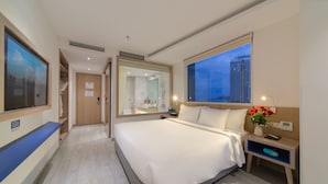 Habitación de hotel con una cama grande, baño con ducha, ventana con vista a la ciudad y escritorio con lámpara y un jarrón de flores.