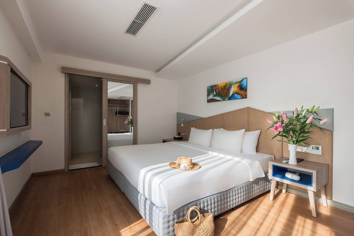 A hotel room with a large bed, a bedside table with a vase of flowers, a wall-mounted TV, and a sliding door leading to another room.
